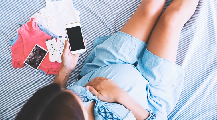 Young pregnant woman with smart phone in hand lying at bed with baby clothes and ultrasound image.